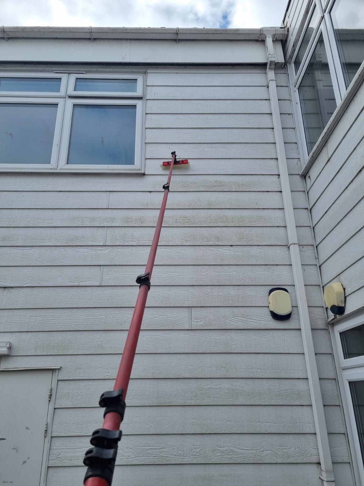 Person using a long red pole with a brush to clean the side of a building with white siding and windows.