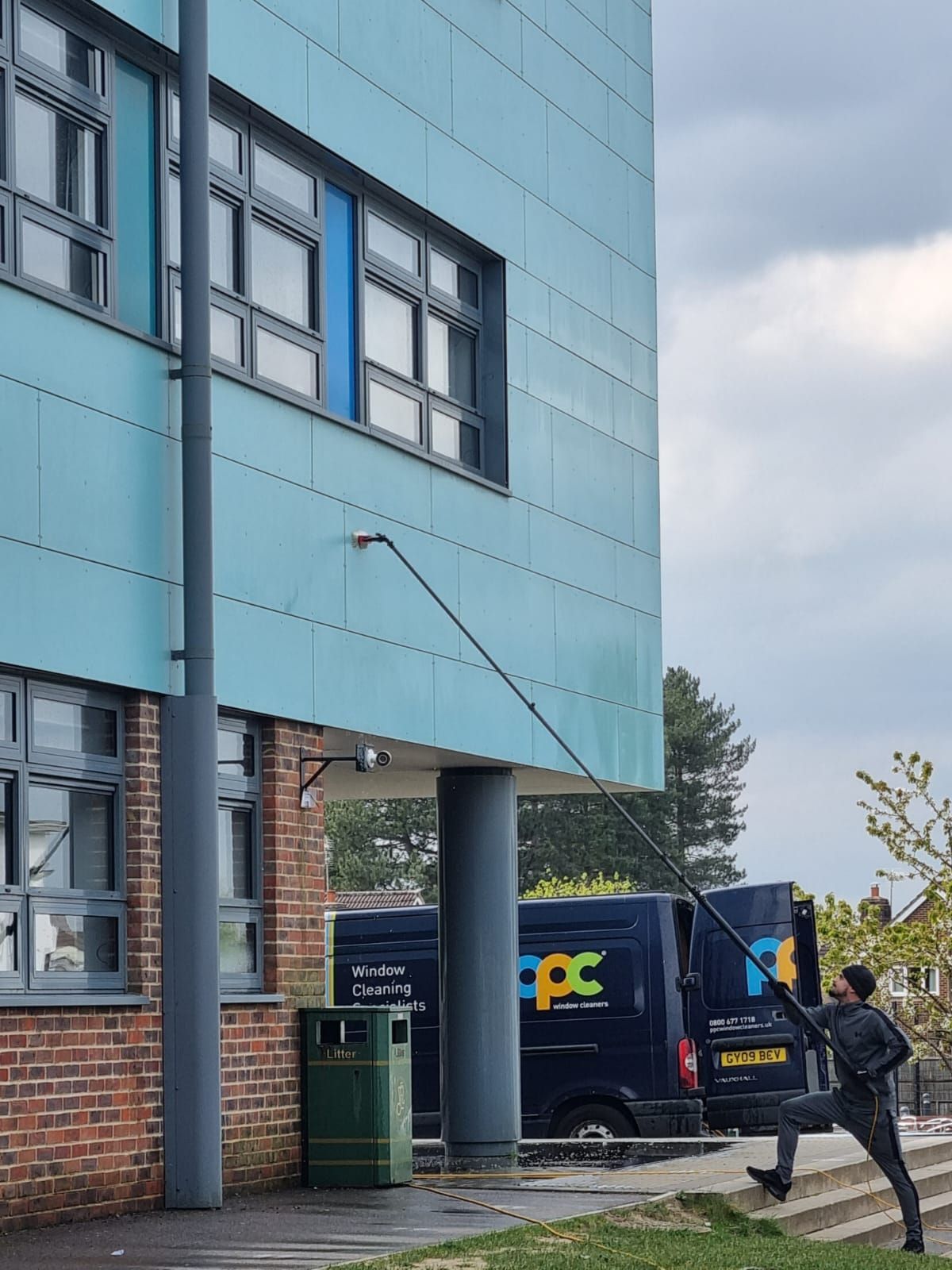 A man washing a window on a turquoise building with a long pole, near a van and trash can.