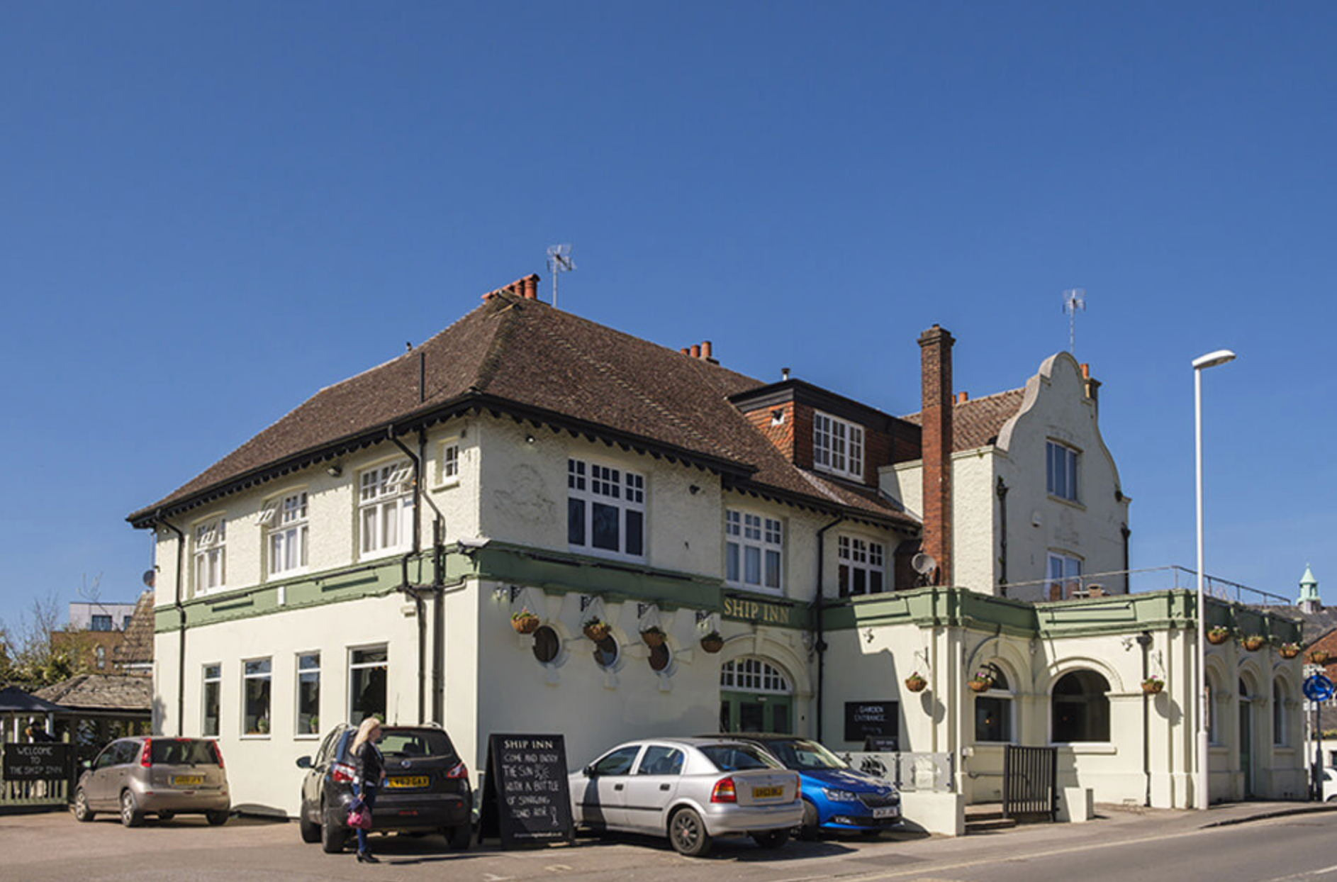 A two-story pub with cream walls, a brown roof, and parked cars out front under a blue sky.
