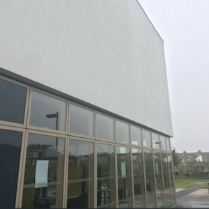 White building with a long row of windows at its base, reflecting a cloudy sky and greenery.