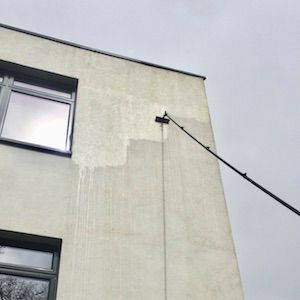 A person uses a pole to wash a light-colored building exterior, leaving water streaks and a clean patch.