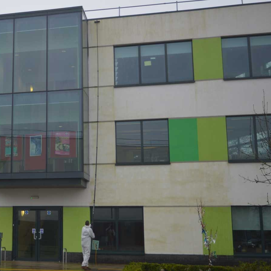 Person in white suit cleans building facade with a long tool; green and white panels, glass windows.