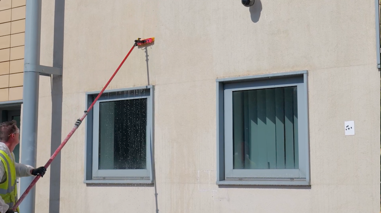 Person washes building windows with a long-handled brush, water cascading down the wall.