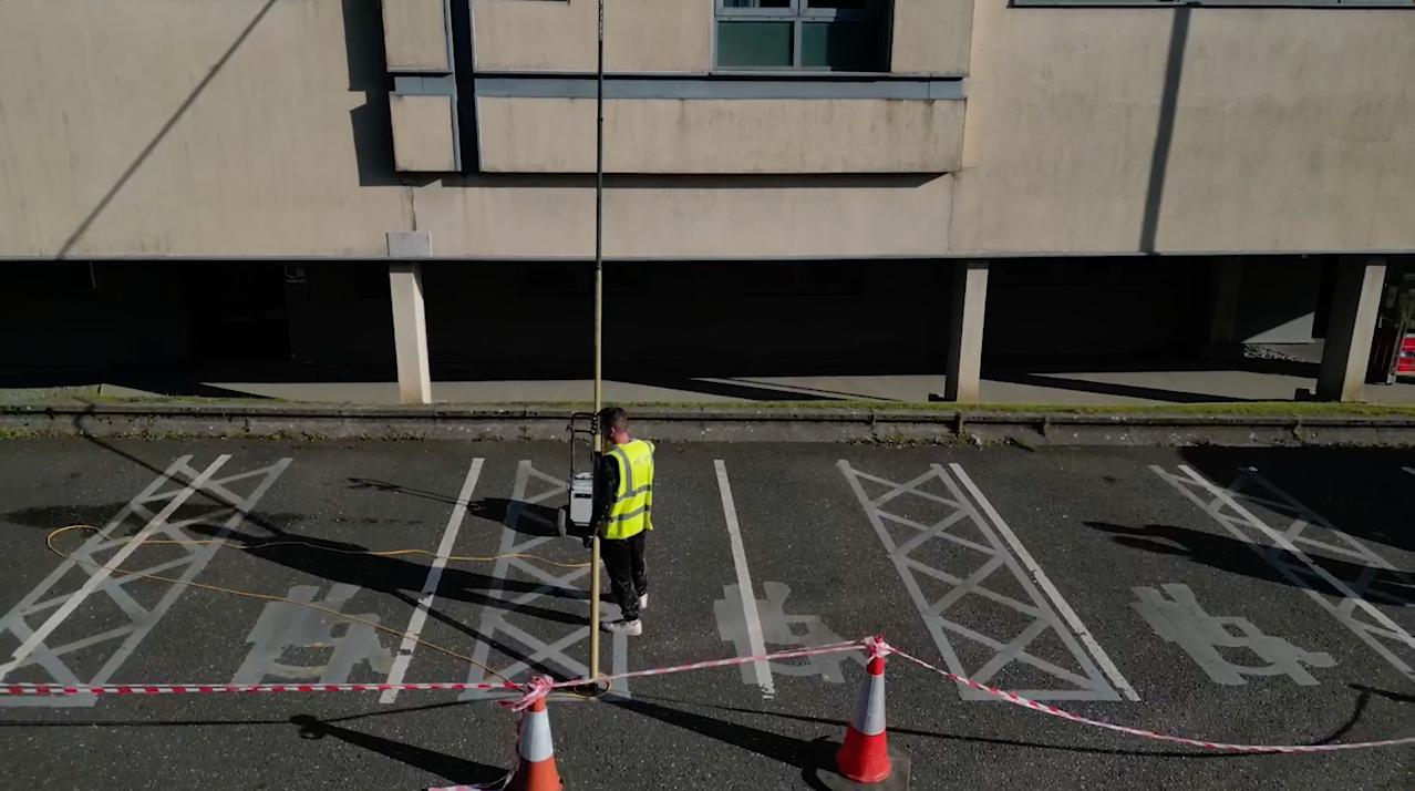 Person in safety vest using surveying equipment in a parking lot with disability parking symbols and road markings in front of a building.