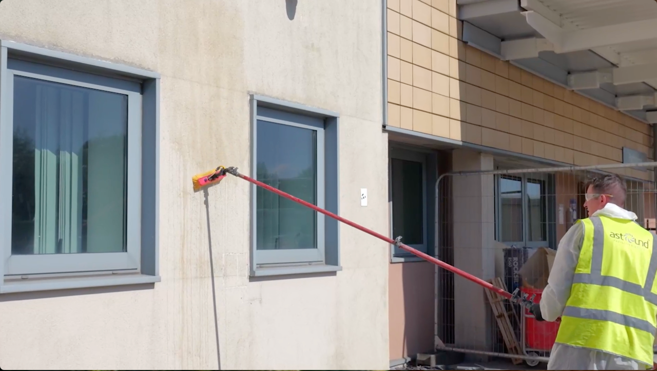 Man in yellow vest washing building exterior with a long-handled tool.