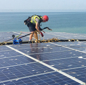 A man in a red helmet cleans solar panels on a rooftop, the ocean in the background.