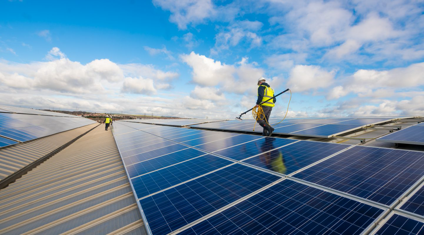 Worker cleaning solar panels on a rooftop against a blue sky with clouds.