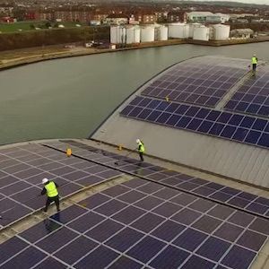 Workers cleaning solar panels on a curved roof overlooking water and industrial tanks.