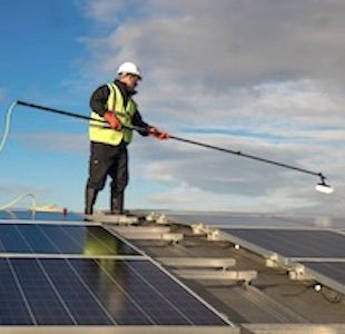 Man in safety gear cleaning solar panels with a long brush on a rooftop.