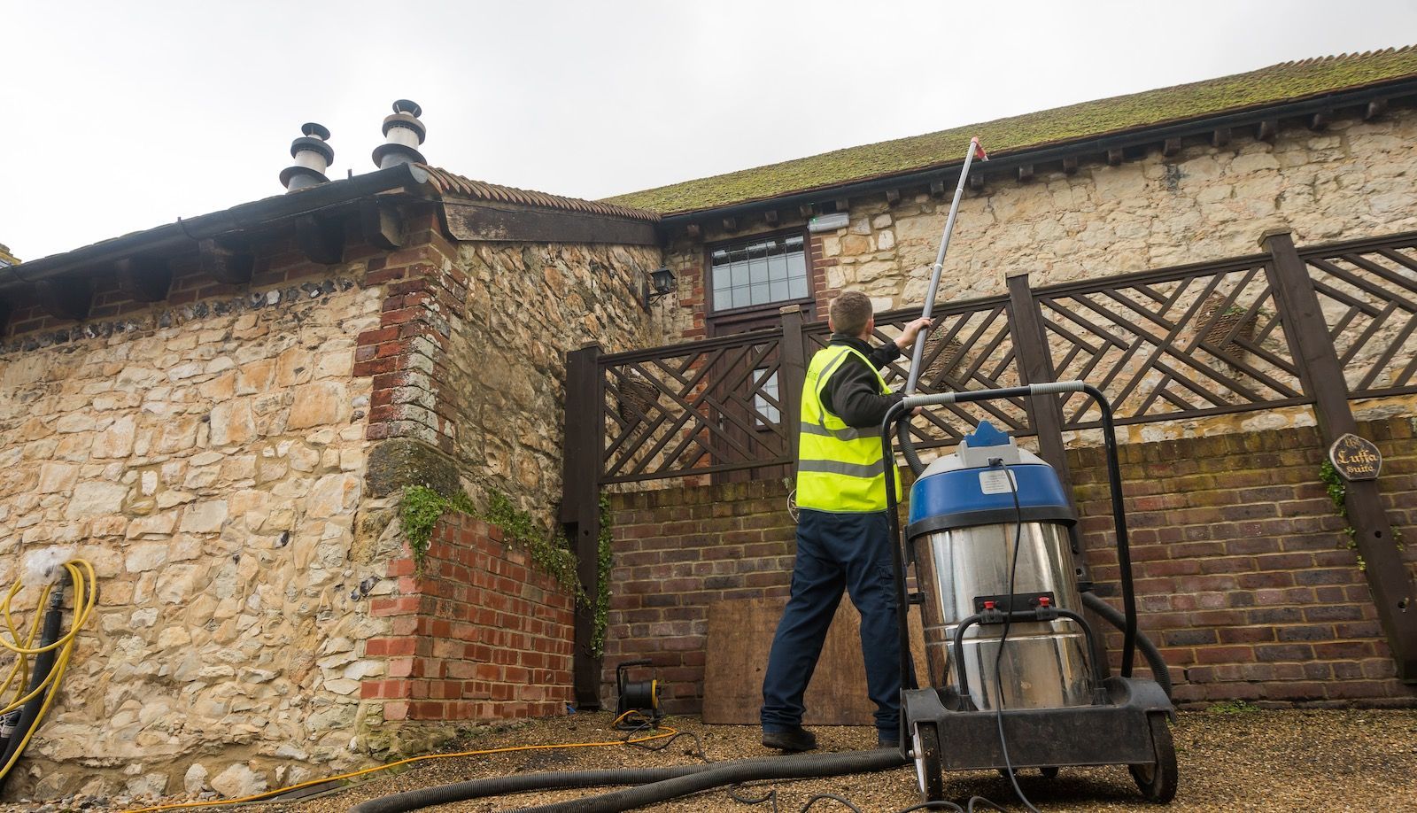 Person in safety vest vacuums a building's exterior with a commercial machine; cloudy day.