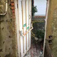 A white door propped open with a metal bar. Brick and weathered walls, a small outdoor area visible beyond the door.