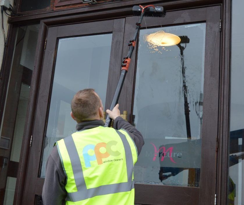 Man in yellow vest cleaning windows with a long pole; outdoors near a building.
