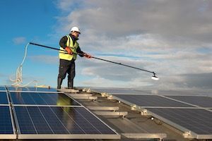 Person cleaning solar panels on a rooftop, using a long brush. Blue sky, safety vest, helmet.