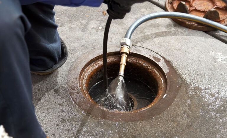 Uma mangueira enche com água uma abertura de drenagem metálica redonda em um piso de concreto.