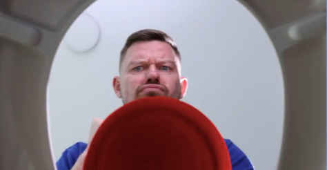 Homem olhando para dentro de um vaso sanitário, visto de dentro do vaso, com um desentupidor vermelho em primeiro plano.