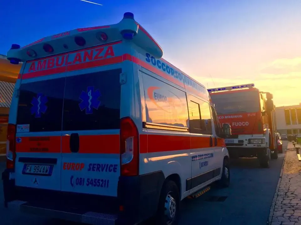 Un'ambulanza parcheggiata a lato della strada al tramonto, con un cielo arancione brillante e un altro veicolo di emergenza davanti.