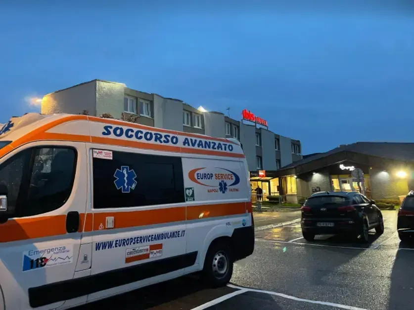 Ambulanza con “soccorso avanzato” parcheggiata fuori da un ospedale al crepuscolo, con un’auto nelle vicinanze