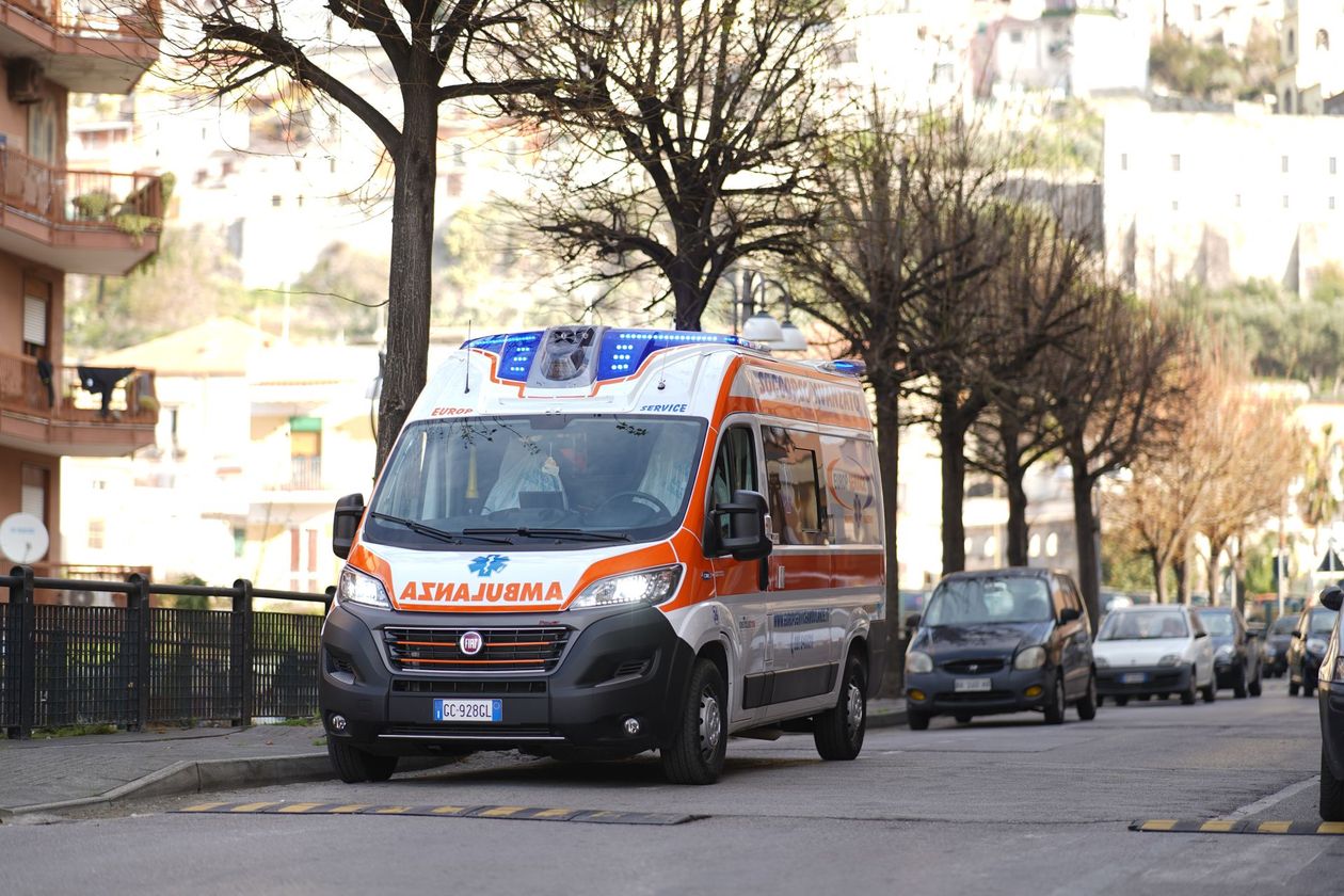 Un'ambulanza su una strada cittadina alberata, accanto ad auto parcheggiate e palazzi residenziali.