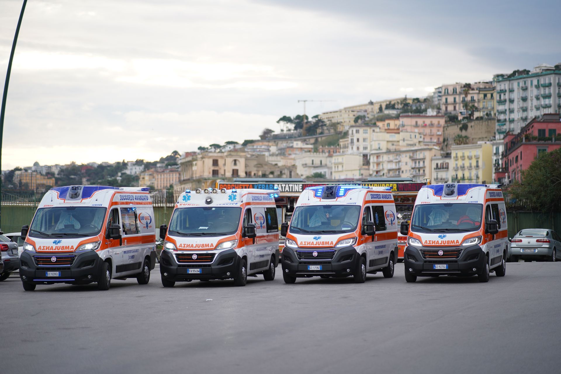 Quattro ambulanze parcheggiate in fila su una strada con edifici sulla collina sullo sfondo.