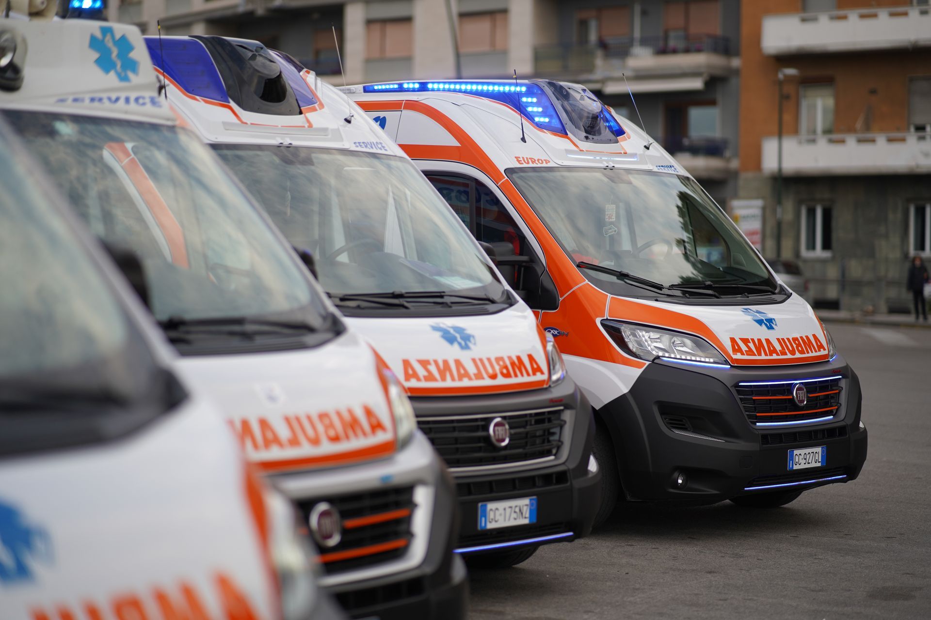 Ambulanze allineate lungo una strada cittadina, con veicoli di emergenza bianchi e arancioni ben visibili.