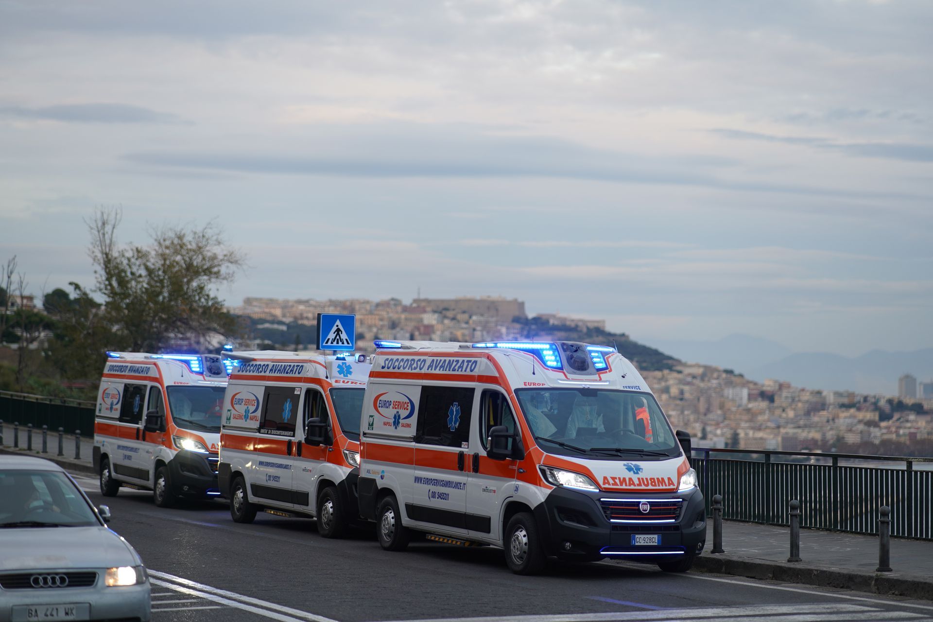 Tre ambulanze con le luci lampeggianti su una strada che si affaccia su una città costiera