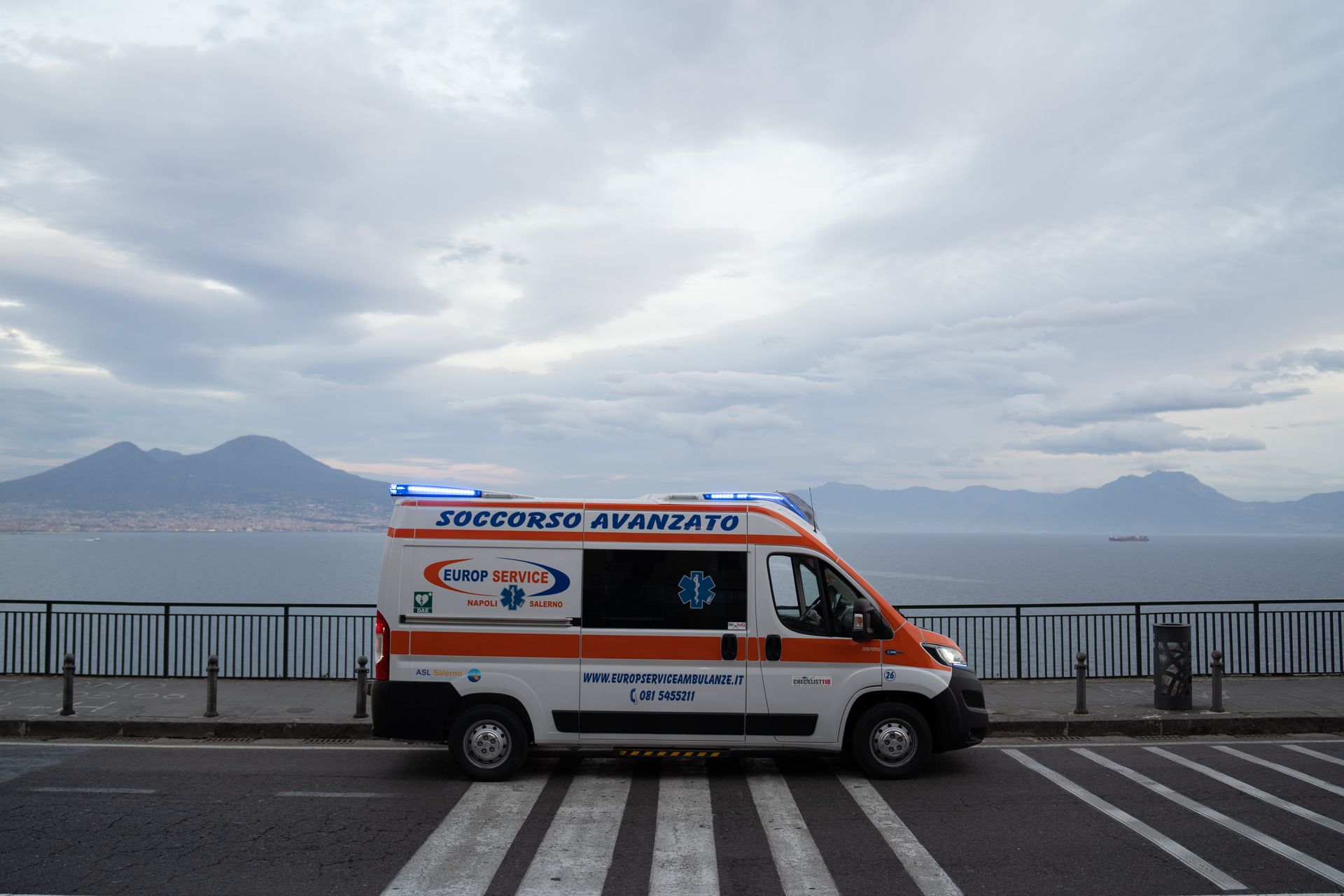 Ambulanza arancione parcheggiata lungo una strada costiera con montagne e cielo nuvoloso sullo sfondo.