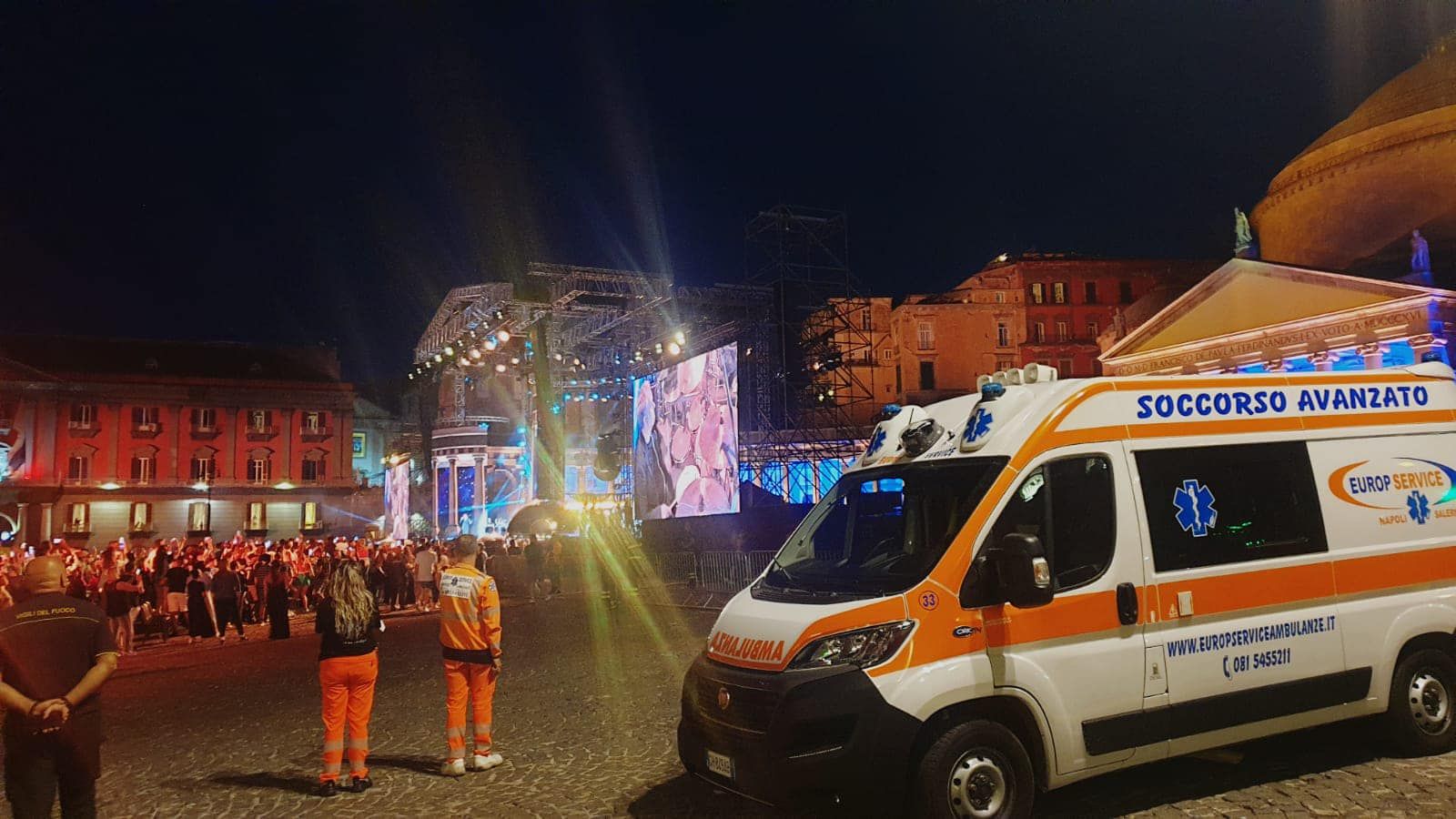 Piazza cittadina notturna con un palco illuminato, folla e un'ambulanza del Soccorso Nazionale in primo piano.