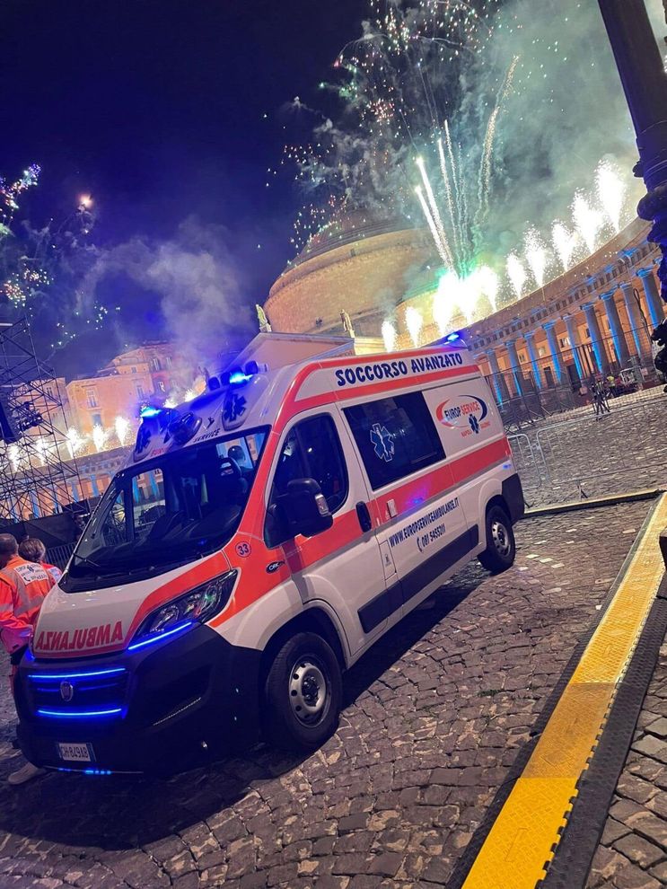 Un'ambulanza con le luci lampeggianti su una strada acciottolata di notte, con fuochi d'artificio e luci festive sullo sfondo.