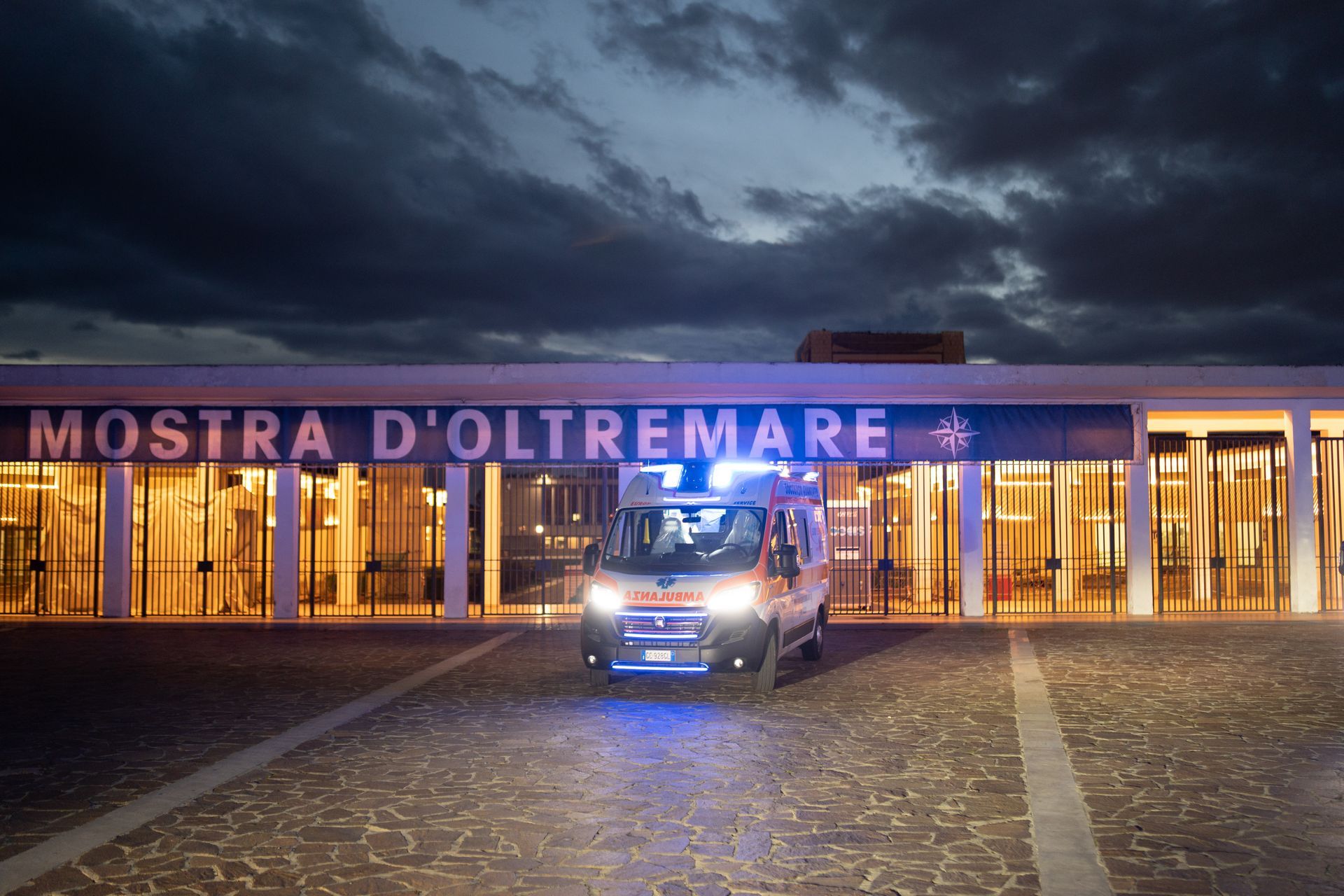 Ambulanza con luci lampeggianti fuori dall'ingresso illuminato della Mostra d'Oltremare di notte