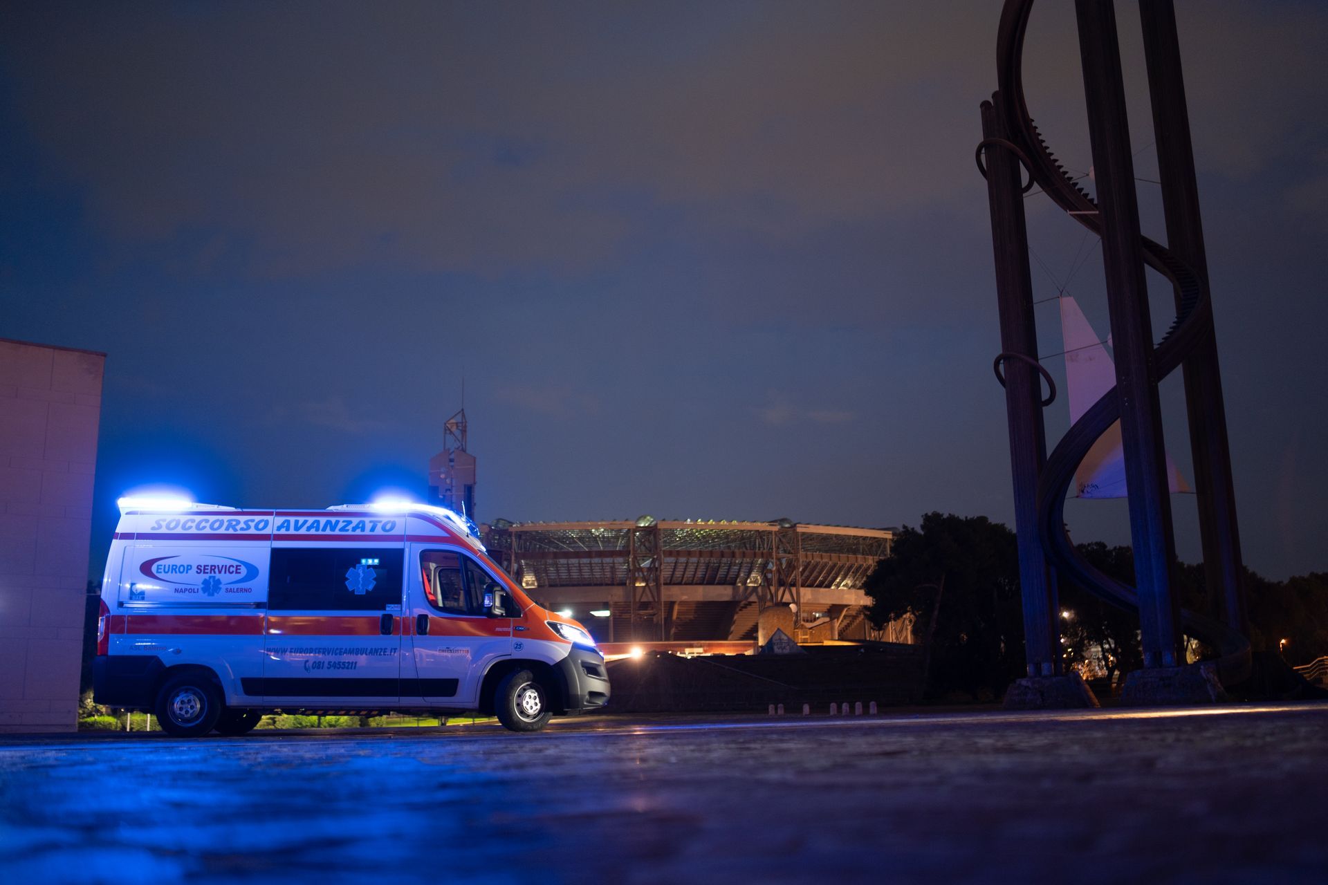 Un'ambulanza con le luci blu lampeggianti è parcheggiata al crepuscolo vicino a un edificio illuminato e a una scultura.