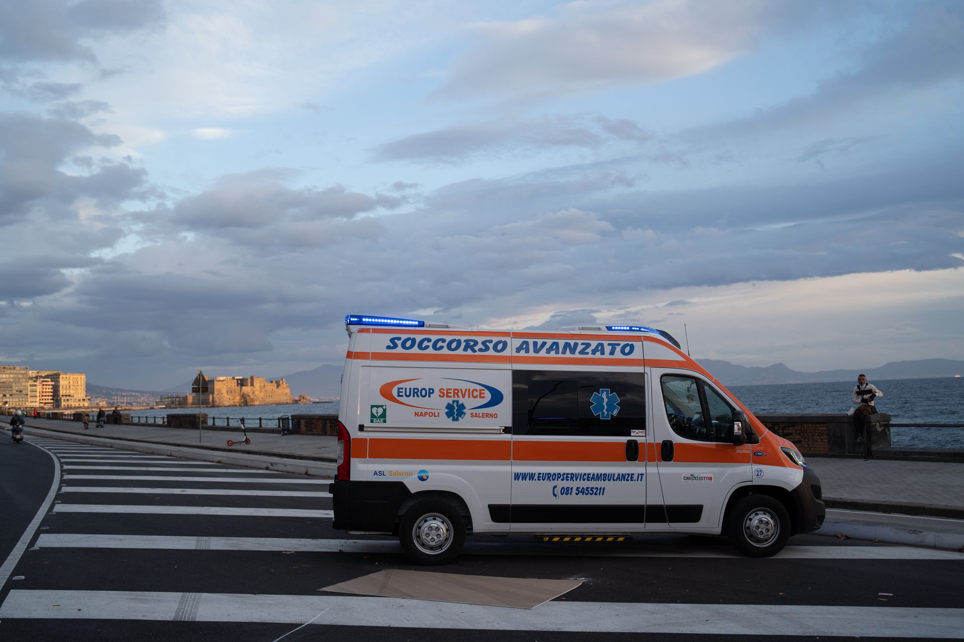 Un furgone ambulanza bianco su un attraversamento pedonale sul lungomare, sotto un cielo nuvoloso.