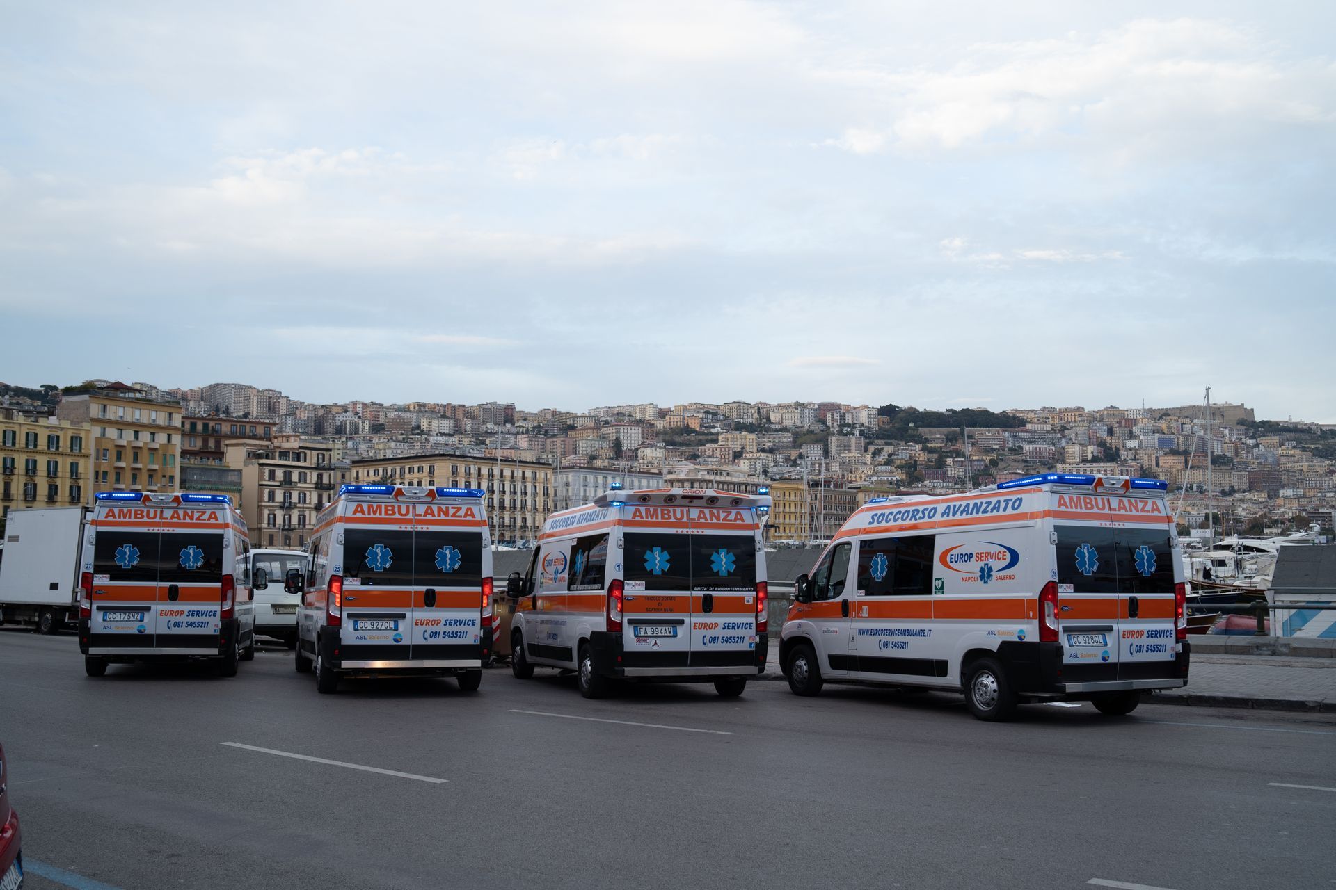 Quattro ambulanze parcheggiate in fila su una strada lungomare con lo skyline della città sullo sfondo.