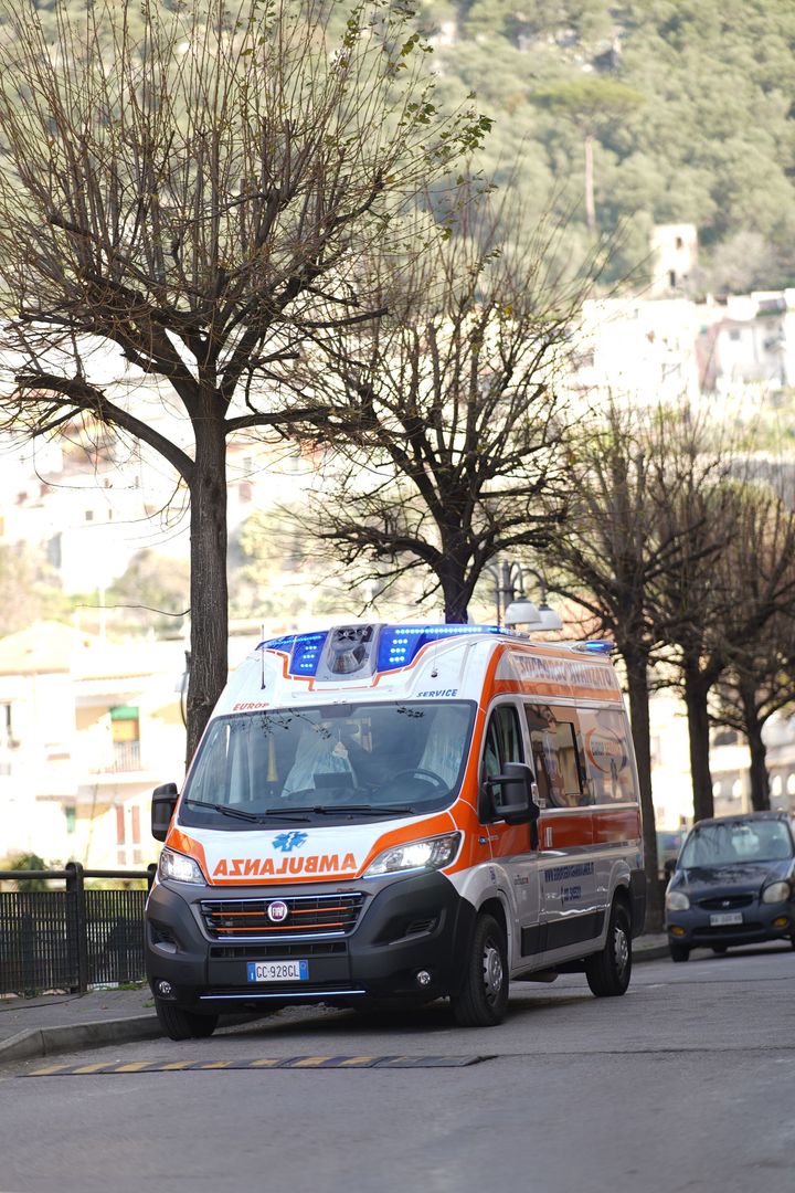 Un furgone ambulanza arancione percorre una strada alberata, con auto parcheggiate e edifici sulla collina sullo sfondo.