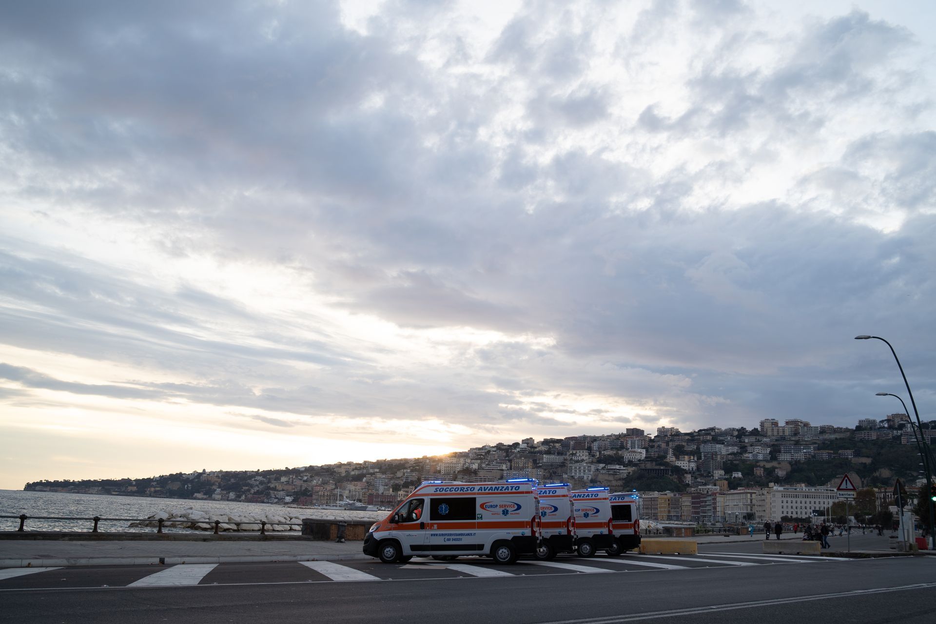 Un'ambulanza parcheggiata su una strada lungomare al tramonto, con colline e cielo nuvoloso sullo sfondo.