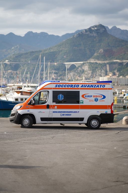 Furgone arancione e bianco parcheggiato vicino a un porticciolo turistico con le montagne sullo sfondo.