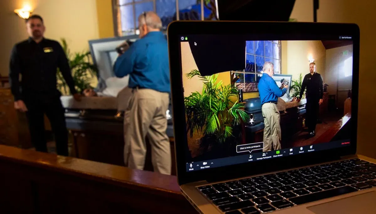 Laptop screen showing two men in a living room, one standing and one adjusting a camera near a plant.
