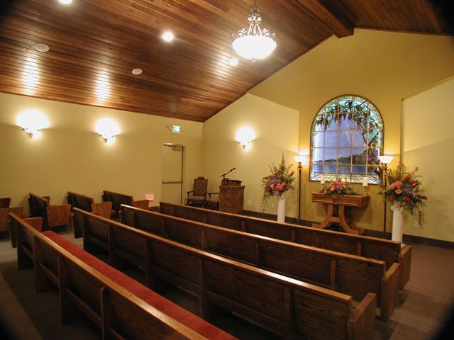 Small church interior with wooden pews, altar, and arched stained-glass window in warm lighting