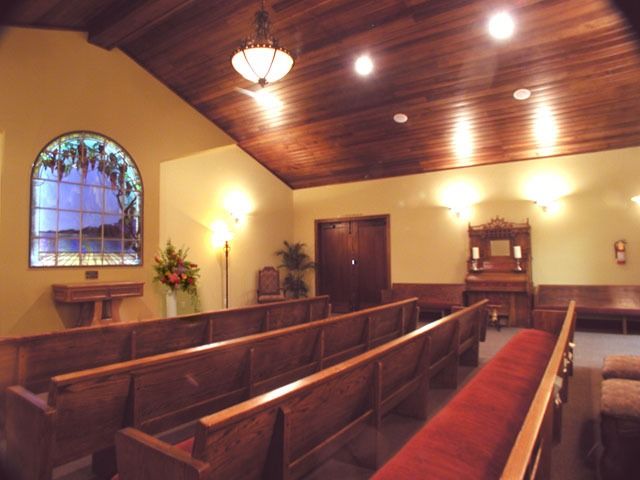Empty church chapel with wooden pews, arched window, and slanted wood-paneled ceiling under warm lights