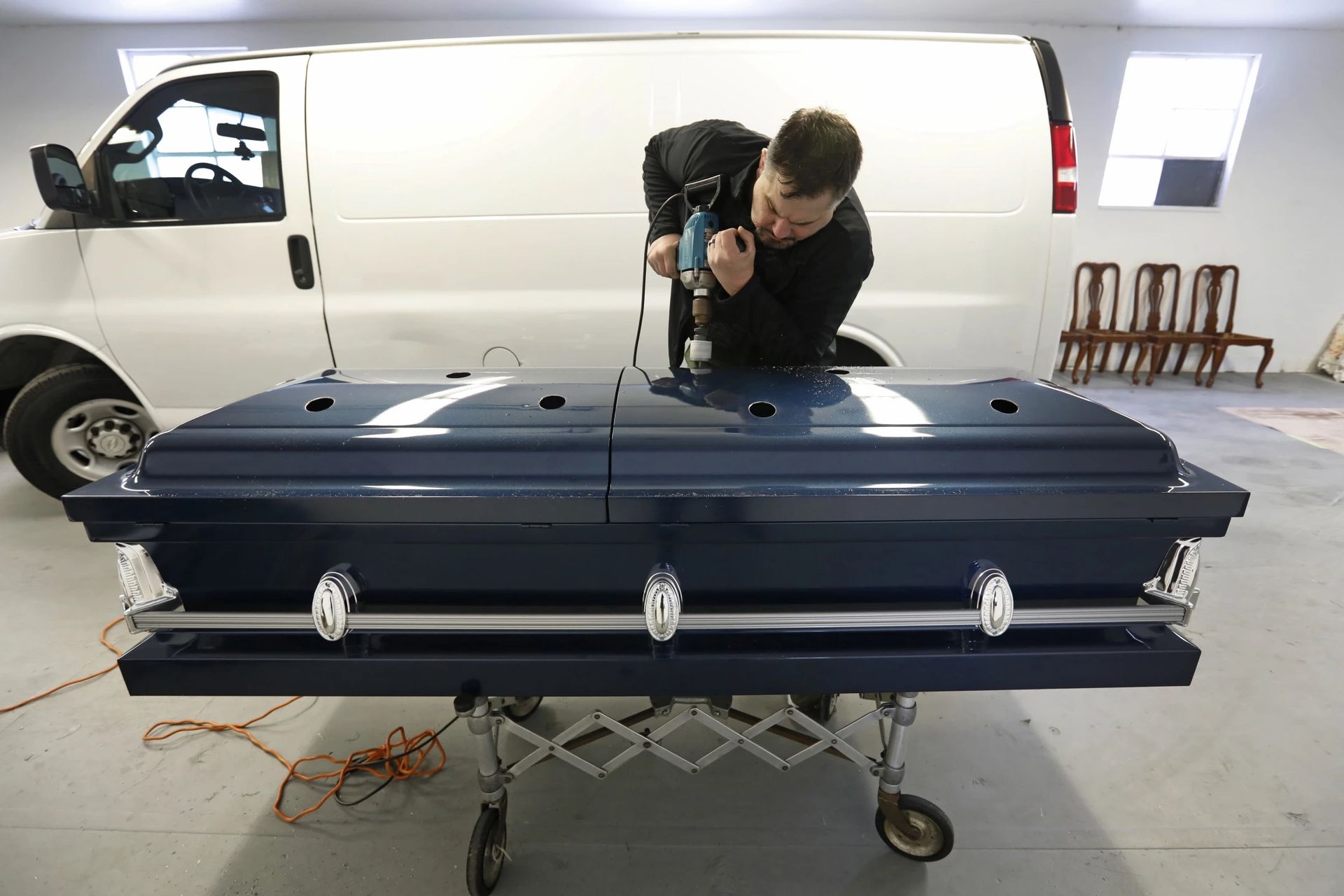 Person photographing a large black coffin on a wheeled stand in a garage, with a white van behind them