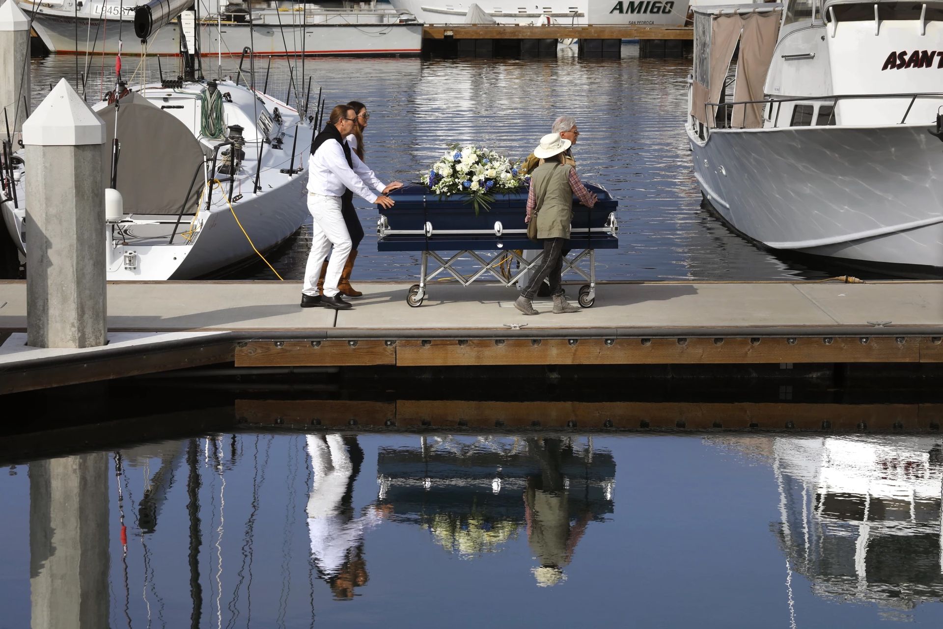 Two people push a blue trolley across a dock beside white boats, reflected in calm water.