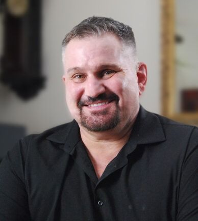 Smiling man in a black shirt seated indoors against a softly blurred background