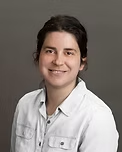 Woman with brown hair and a light blue button-down shirt smiles against a gray background.
