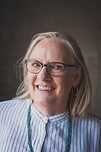 Woman with blonde hair and glasses smiling, wearing a striped shirt and turquoise necklace.