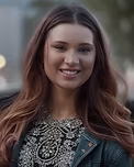 Woman with brown hair smiles, wearing a decorated top and denim jacket; outdoor setting.