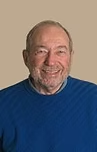 Smiling older man with a white beard, wearing a blue sweater, against a neutral background.