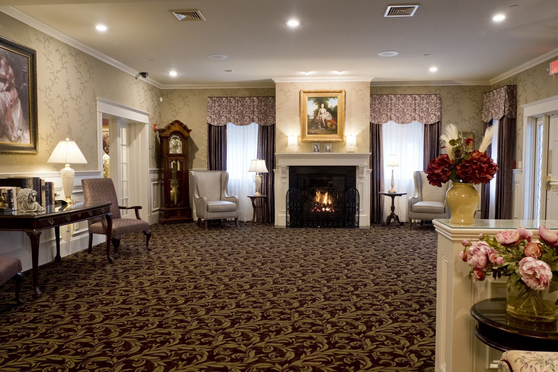 Interior of a room with fireplace, chairs, flowers, and a grandfather clock.