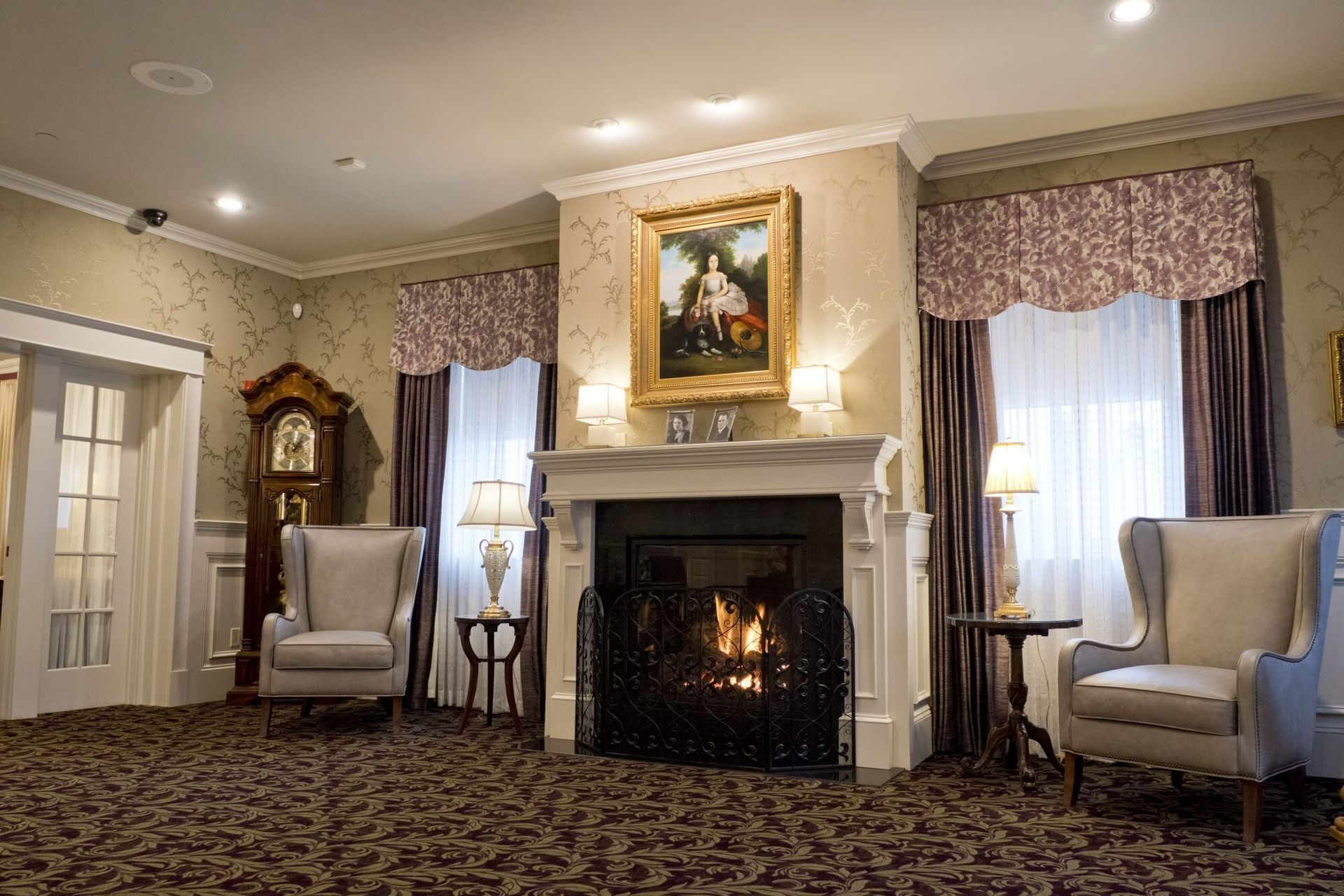 Cozy room with fireplace, chairs, grandfather clock, and artwork. Warm colors, ornate decor.