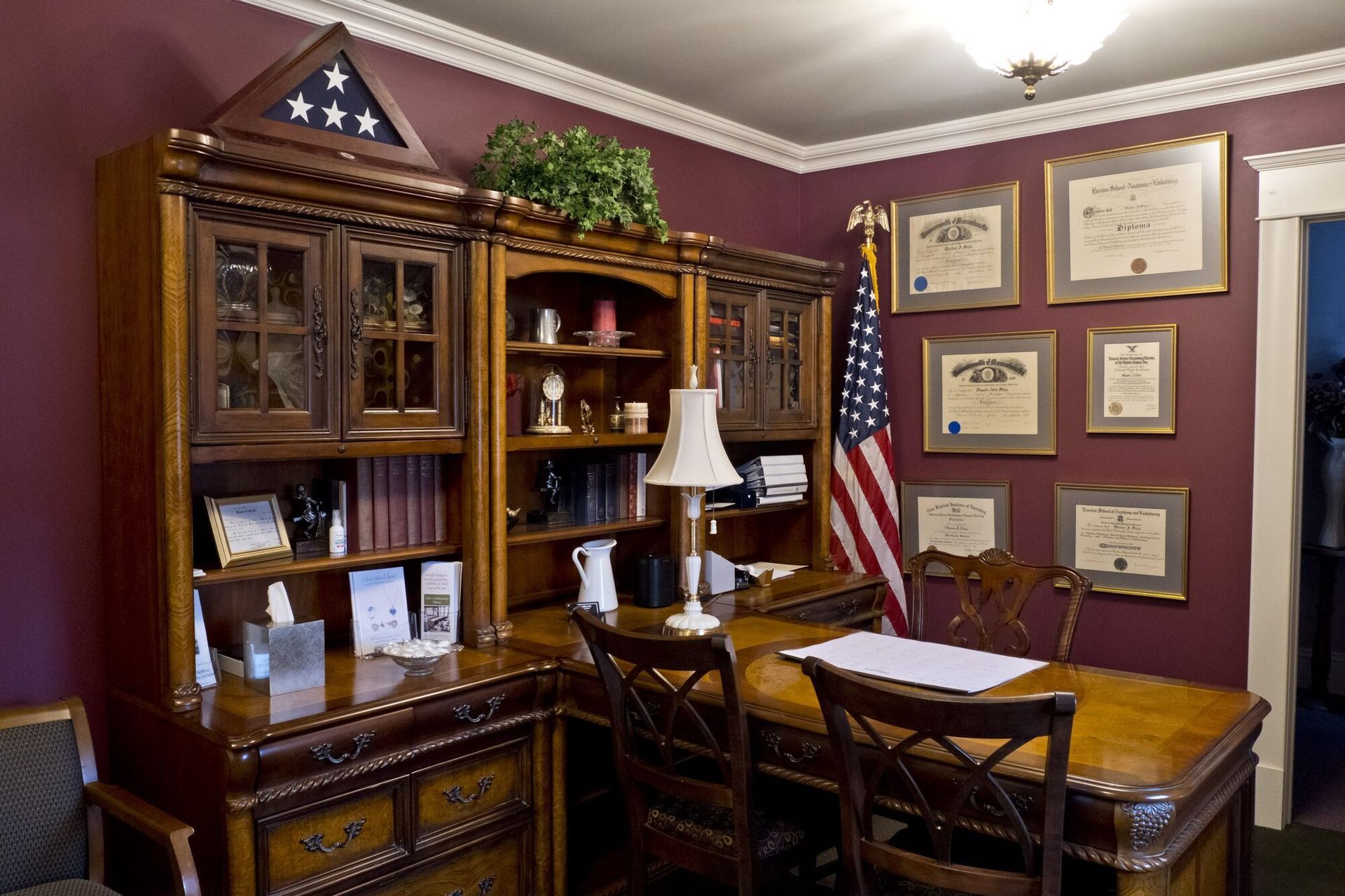 An ornate wooden office with desk, bookshelves, and an American flag. Diplomas on the maroon walls.