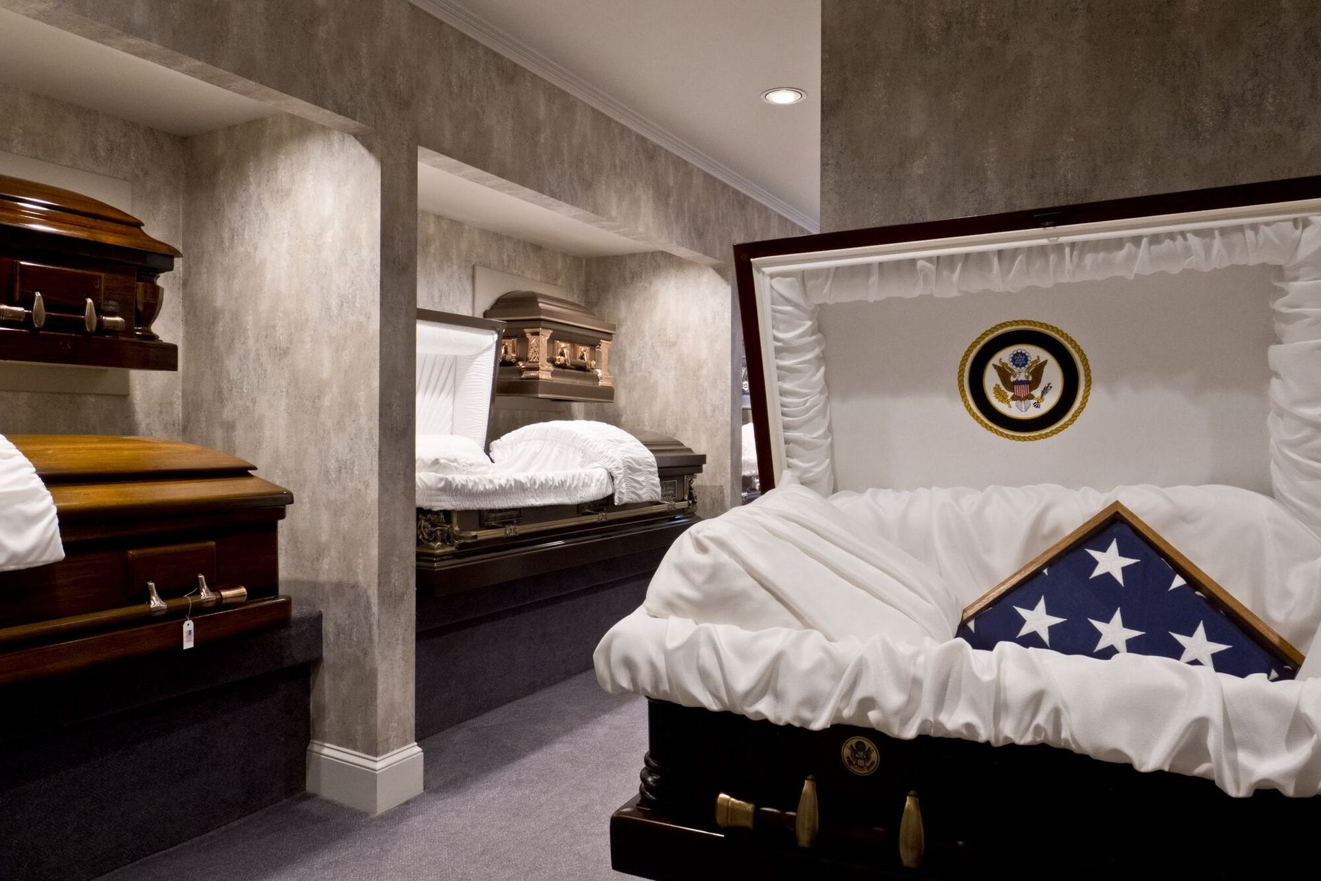 Interior of a funeral home displaying several open and closed coffins. One coffin holds an American flag.