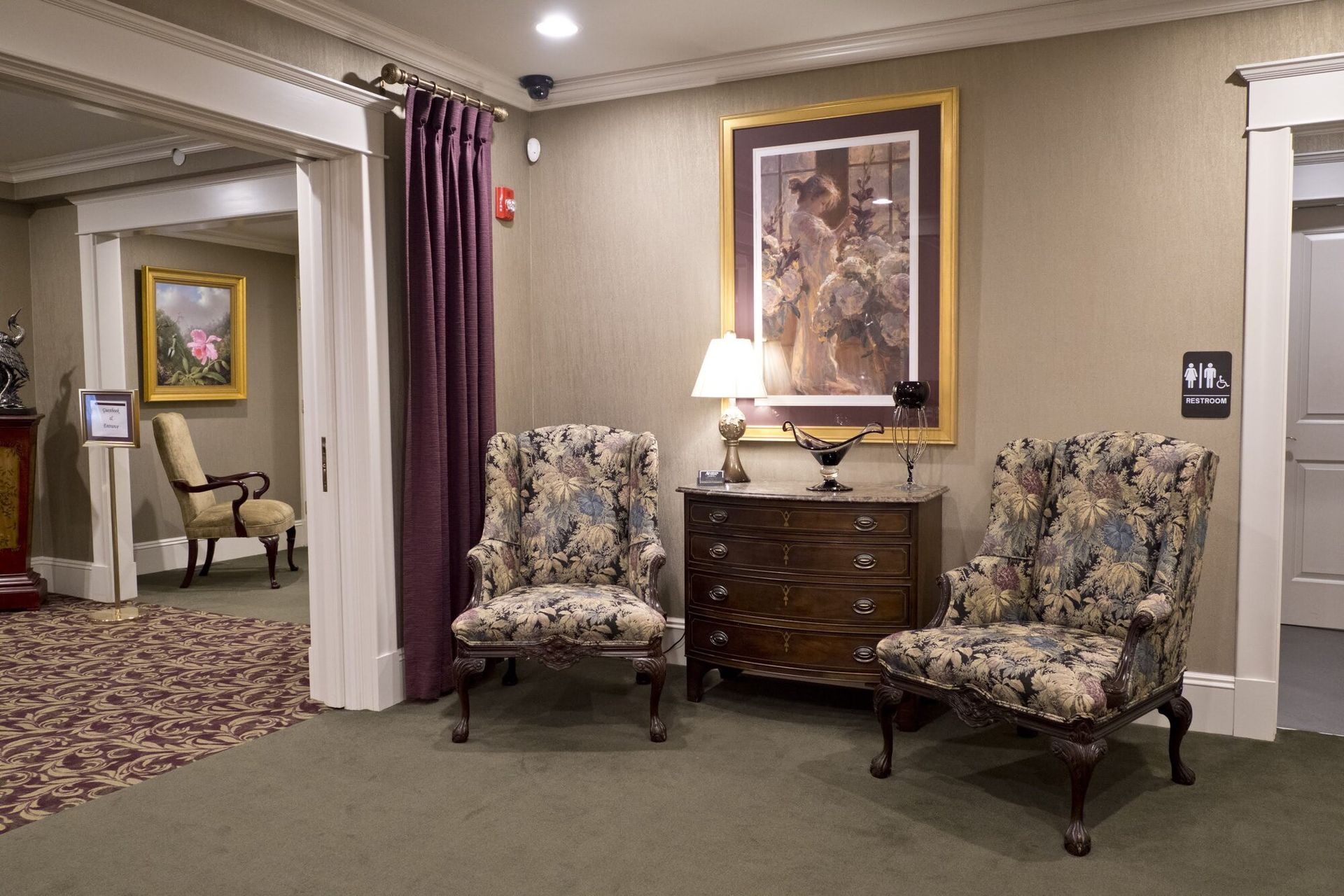 Lobby with two patterned armchairs, a chest, a painting, and a doorway to another room with more seating.